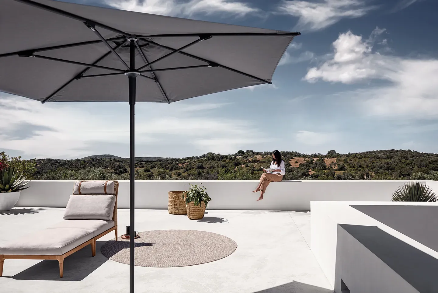 one halo push up parasol and a lima lounger with deco rug on a rooftop terrace with a woman seated on a wall and landscape behind ff