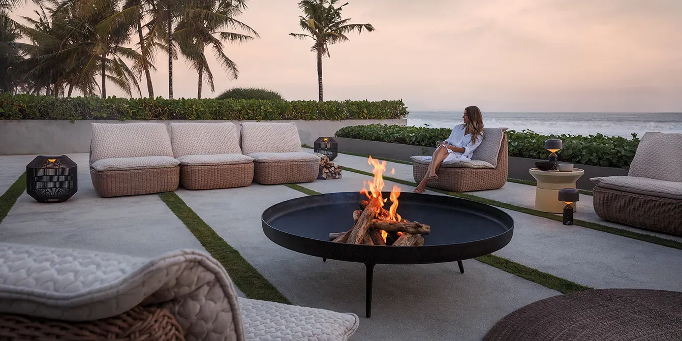 a woman sits on an omada lounge seating unit amongst a large omada seating group with a deco fire bowl ambient nest and ambient pebble lanterns and a coso table with a tropical sunset behind
