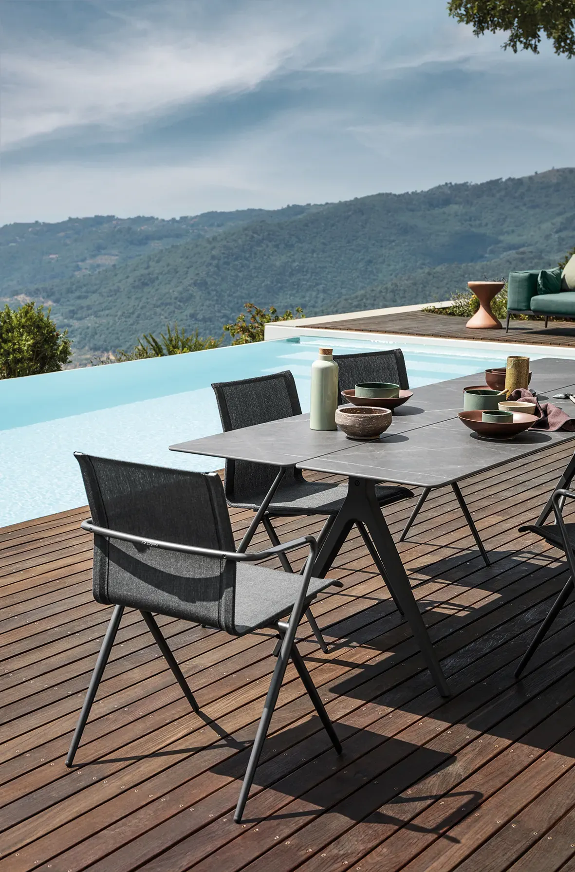 3 ryder dining chairs surround a split dining table with ceramic top on a poolside deck with forest and sky behind