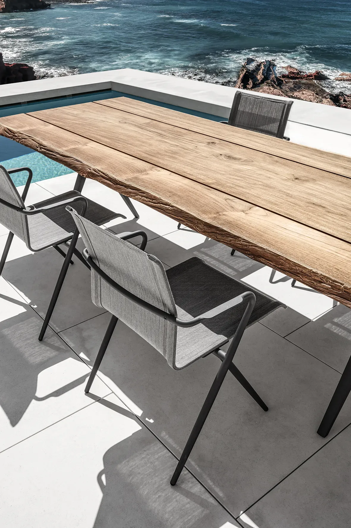 ryder dining chairs surround a split dining table with raw teak top on a poolside terrace overlooking the sea