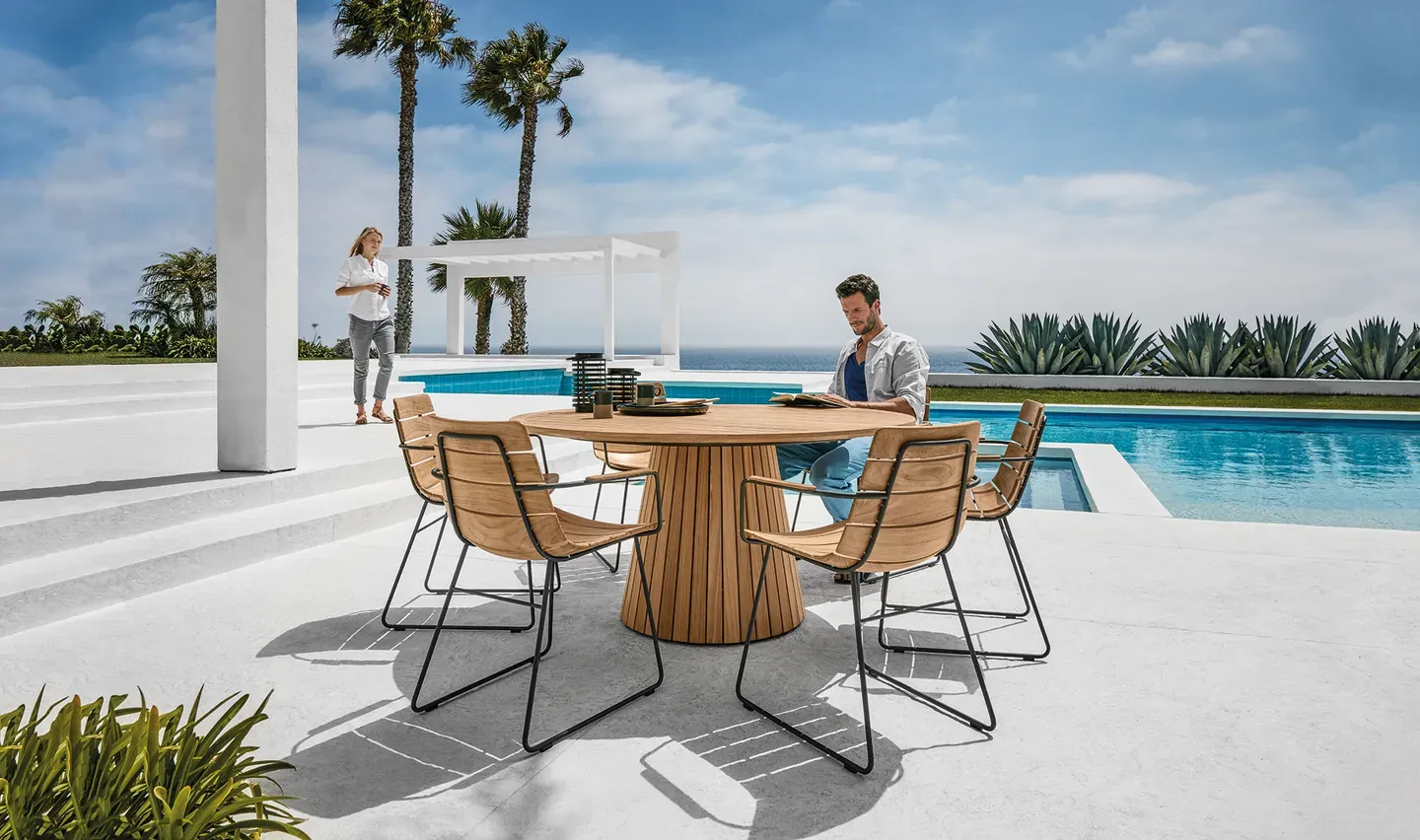 a woman walks behind a man seated at a whirl teak dining table with william dining chairs by a pool with blue sky